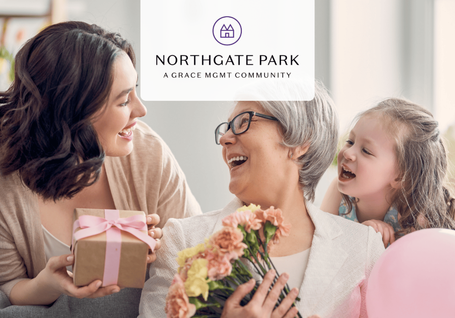 Three generations sharing a joyful moment: a younger woman presents a pink‑ribboned gift to an older woman holding flowers, while a young girl smiles beside them with a balloon nearby.