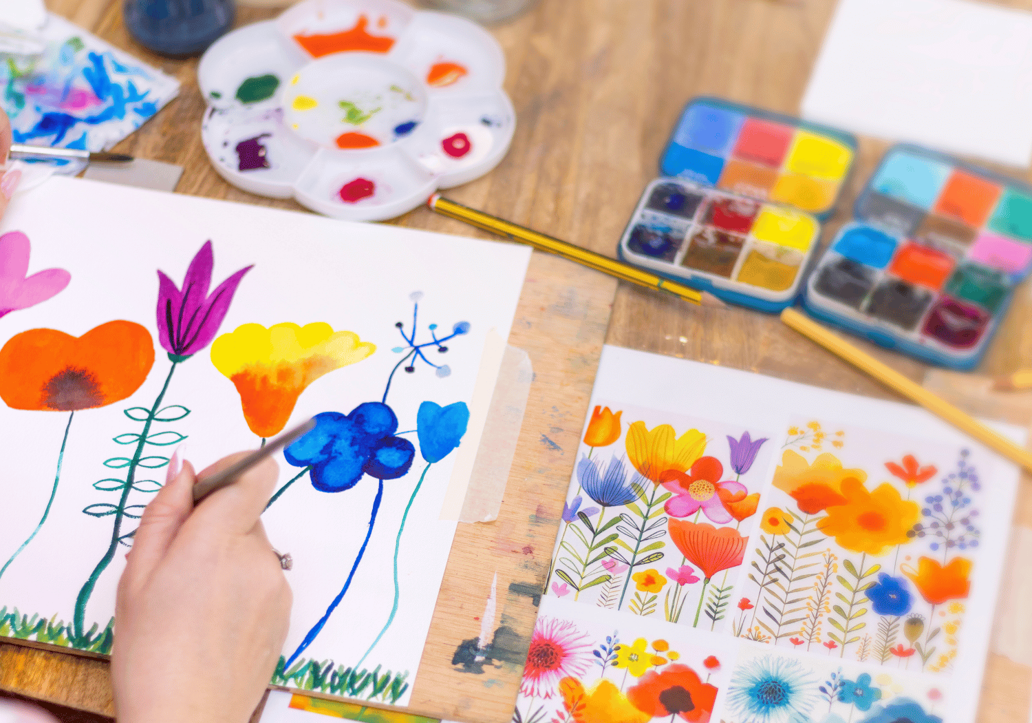 A person paints vibrant watercolor flowers on white paper, with color palettes and brushes on a wooden table nearby.