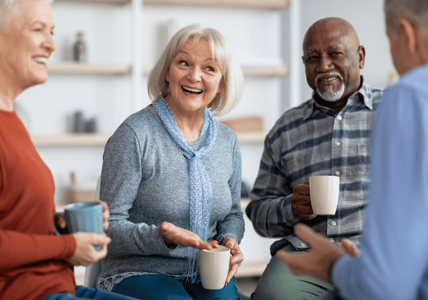Group of three seniors smiling and talking, each holding a mug in a bright, cozy room.